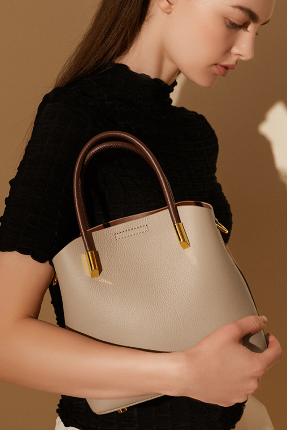 Woman holding a beige handbag with gold accents against a brown background